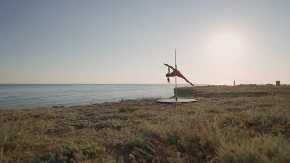 Athletic Woman in a Pink Tracksuit Performs Tricks on a Pole on Background Sea