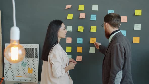 Cheerful Colleagues Man and Woman Talking Looking at Sticky Notes on Wall in Office