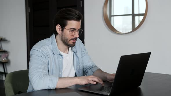 A young Man with Glasses, a Blue Shirt and a White t-Shirt is Working on a Computer laptop