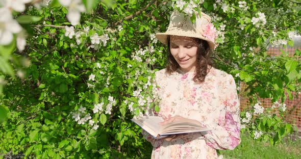 Slow Motion, Girl in Hat Sitting in Park Near a Flowering Tree Is Reading Book. Close-up, View of