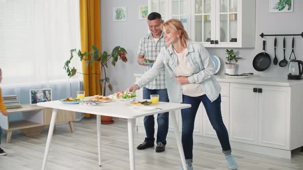 Happy Married Couple Together with Their Son Put Plates of Food on Table for Dinner Together at Home