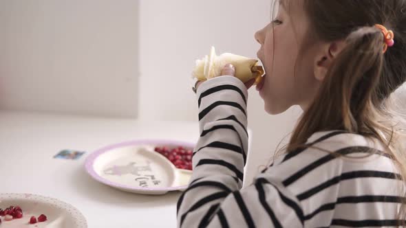 A Little Girl is Sitting on Chair and Eats Thin Pancakes in Striped Shirt