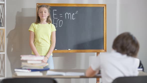 Confident Intelligent Caucasian Schoolgirl Writing Physical Formulas on Chalkboard in Classroom