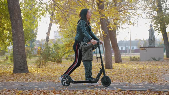 Young Mother Showing Her Toddler Son How To Ride a Scooter in a Autumn Park. Active Family Leisure