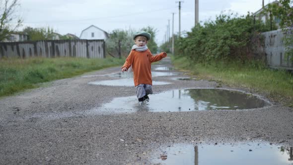 Outdoor Games, Lovable Cheerful Boy in Hat and Rubber Boots Plays in Puddles on Street After Rain