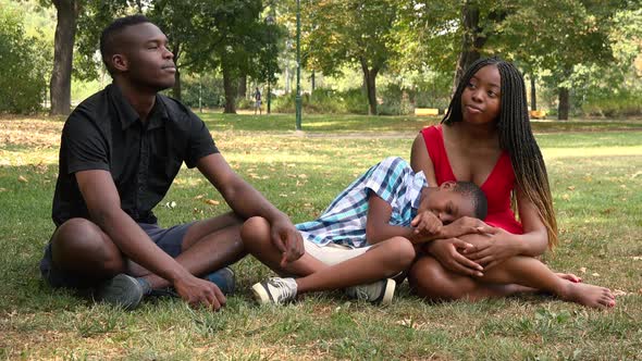 A Young Black Family Sits on Grass in a Park, a Boy Lies with His Head on His Mother's Lap