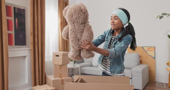 Girl Helps Her Parents Unpack Boxes After Moving Finds Cardboard Box with Her Toys Pulls Out Teddy