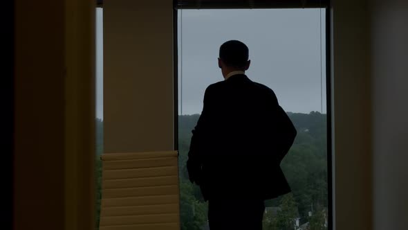 Businessman in a Black Suit is Walking Towards a Big Window That Looks at the Buildings