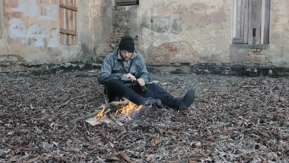 Homeless Man Sits on Ground and Pushes Pieces of Cardboard Into Fire to Keep Warm From Cold