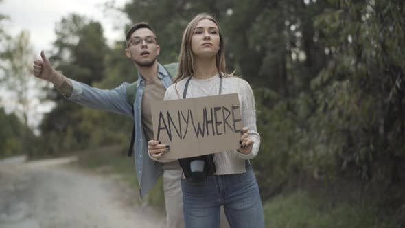 Positive Millennial Caucasian Couple Holding Anywhere Sign and Hitchhiking on Roadside