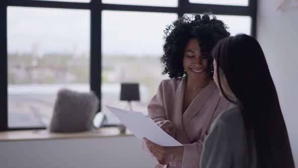 Positive African American Woman Giving Highfive to Asian Friend Looking Through Paperwork Indoors