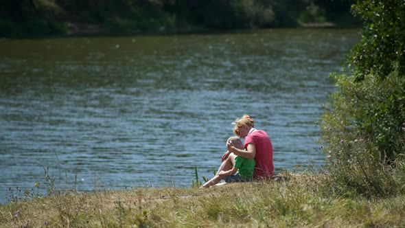 A young mother and a little boy sit on the Bank of the river . Mom puts on her son's headphones.