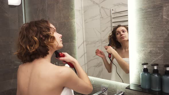 A Young Woman in Big White Bath Towel Styling Her Hair with a Curling Iron in Front of a Mirror