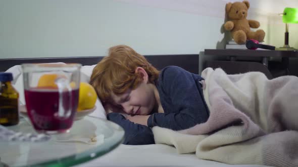 Portrait of Crying Caucasian Little Boy Lying in Bed in Front of Pills, Oranges and Hot Tea Standing