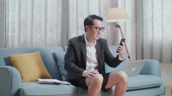 Asian Businessman In Jacket And Shorts Having Video Call On Smartphone While Working With A Laptop