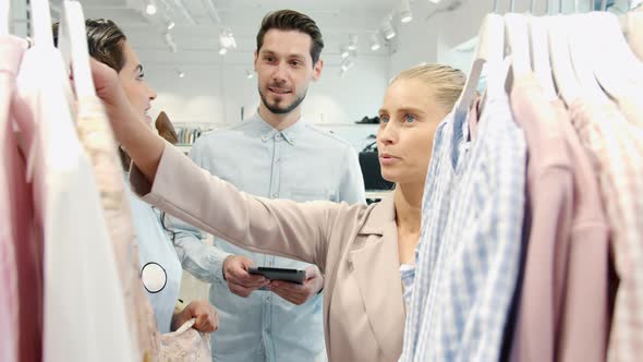 Young Women Friends Choosing Clothes and Talking to Salesman Holding Tablet in Shop