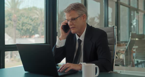 A businessman talking on the phone while working on a laptop at the office