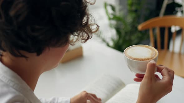 A Beautiful Woman Holds A Cup Of Cappuccino Coffee Sits Reading A Book On A Clear Bright Day In Cafe