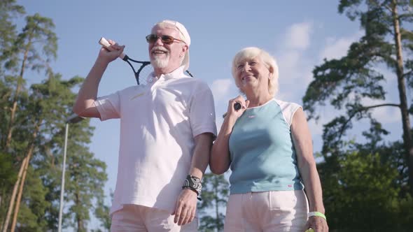 Mature Couple in Sunglasses and Tennis Rackets Standing on a Tennis Court in the Sun