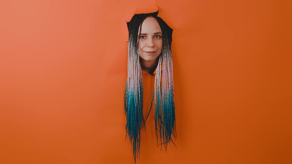 Young Woman with Multicolored Hairstyle Sticking Out of Hole of Orange Background