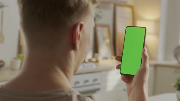 Back View of Young Man at Home Sitting on Kitchen Room Using With Green Mockup Screen Smartphone