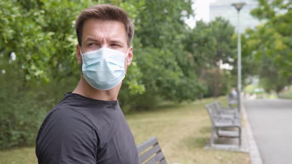 A Young Handsome Man in a Face Mask Looks Around As He Sits on a Bench in a Park in an Urban Area