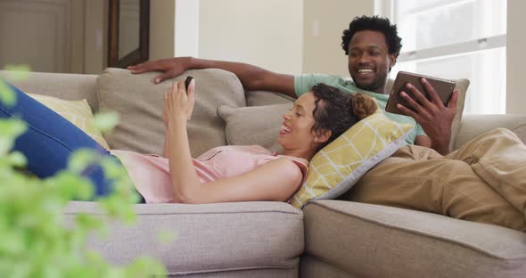 Happy biracial couple relaxing on sofa with smartphone and tablet