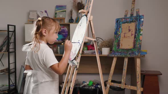 Beautiful Concentrated Little Girl Paints with Paintbrush on Canvas on an Easel