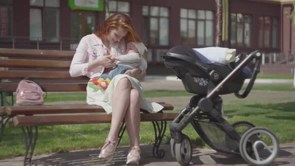 Beautiful Red-haired Woman Playing with Her Child Sitting on the Bench. The Stroller Standing Near