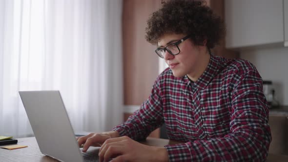 A Curly Man with a Serious Look Works at a Laptop Sitting in a Modern Kitchen