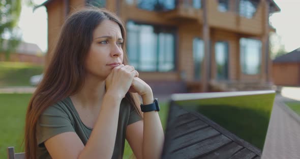 Frustrated Young Woman Sitting in Front of Laptop at Table in Courtyard of the House