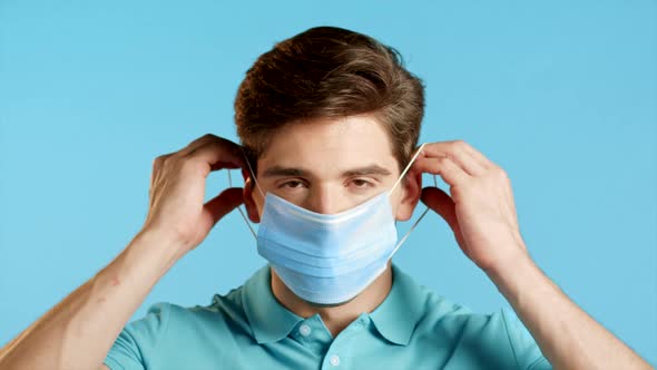 Young Man Puts on Face Medical Mask During Coronavirus Pandemic. Portrait on Blue Background