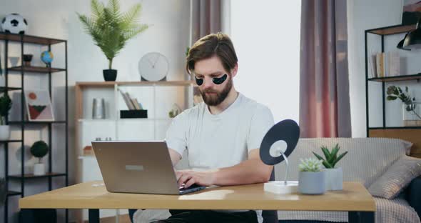 30-Aged Bearder with Anti-Aging Hydrogel Eye-Patches Sitting at the Table and Working on Computer