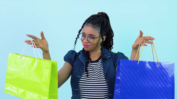 Cheerful Young African American Girl in Blue Clothes Holding Laminated Paper Bags with Pens