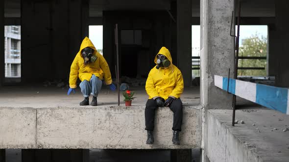 Man and Woman are Sitting on Edge of Abandoned Building in Gas Masks and Yellow Protective Suits in