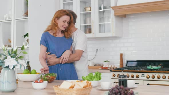 Aged Man Hugging His Attractive Wife in Kitchen