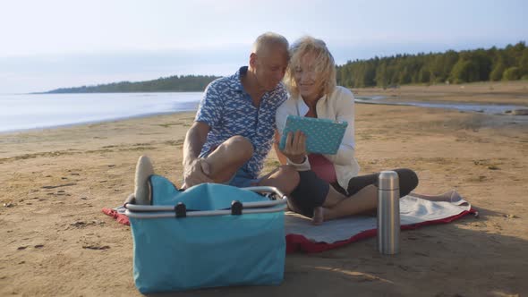 Retired Couple Relaxing on Beach Sitting on Blanket and Using Digital Tablet
