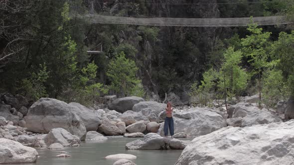 A Woman in Wide Light Pants Does Meditative Exercises on the Rock in the Middle of the Stream