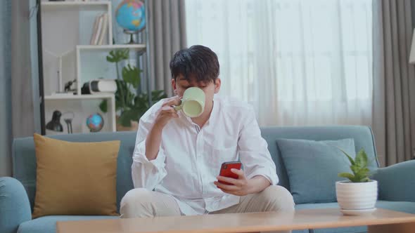 Happy Asian Man Drinking Coffee While Using Smartphone In The Living Room