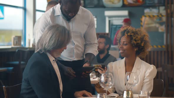 Waiter Showing Menu on Digital Tablet To Multiethnic Couple in Restaurant