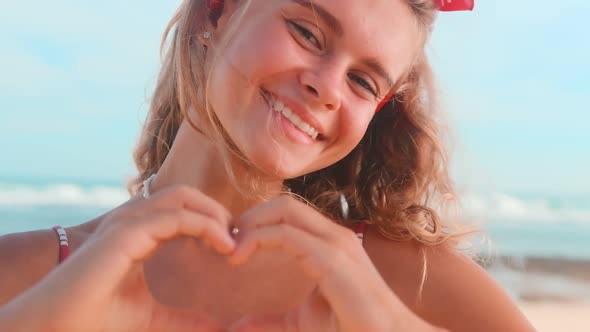 Young Blonde Caucasian Woman Teenager Shows Heart with Fingers Stands on Beach