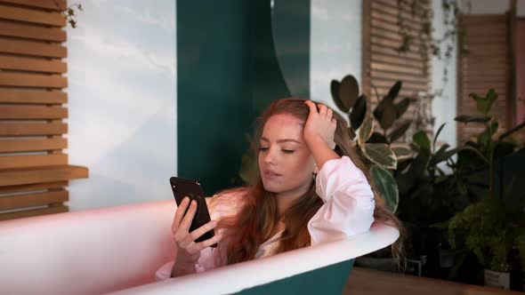 Pretty Girl With Long Hair In White Men's Shirt Sits In The Bathtub In The Bathroom.