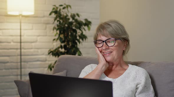 Pretty Middle Aged Woman On Sofa With Laptop Communicates With Someone Via Video Call. She Touches