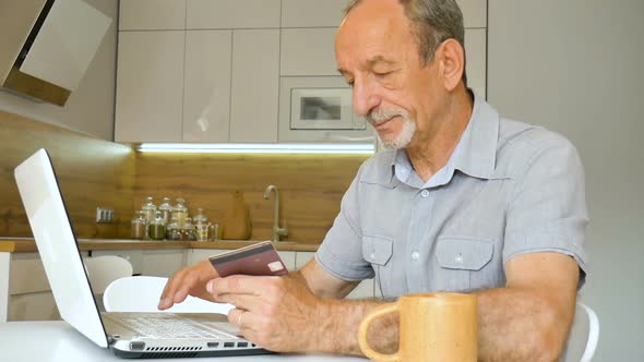 Trendy Mature Man is Working From Home with Laptop Sitting at the Table in His Kitchen Buying