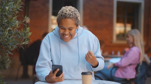 Excited African Woman Receiving Good Email on Smartphone and Making Winner Gesture Sitting in Cafe