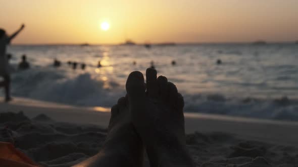 POV Silhouette of Feet of Young Man Lying on Sandy Beach By Ocean During Sunset