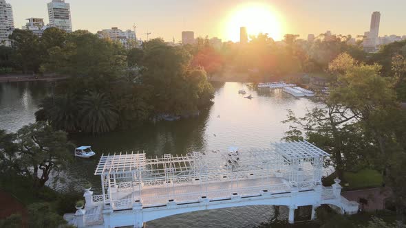 Aerial dolly in over white bridge and boats sailing in pond at Rosedal gardens at golden hour, Bueno