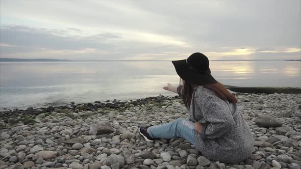 Young Woman Sitting on Pebble Beach Throwing Stones Into the Water