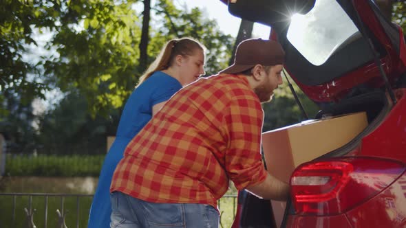 Happy Overweight Couple Taking Cardboard Boxes From Car Trunk on Moving Day