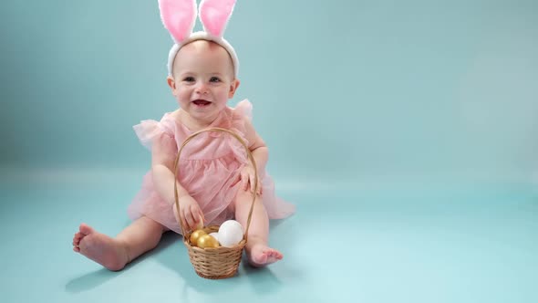 Happy Baby Girl in Bunny Ears Headband Sitting on Blue Studio Background and Playing Easter Eggs